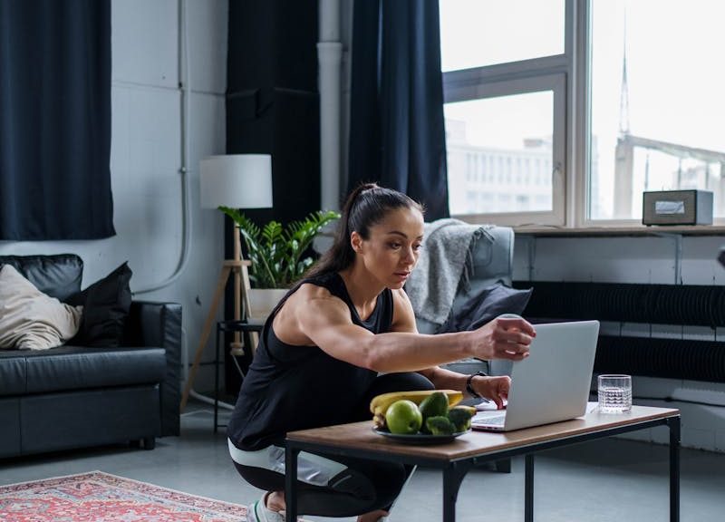 Woman using laptop while exercising at home, featuring a modern gym setting.