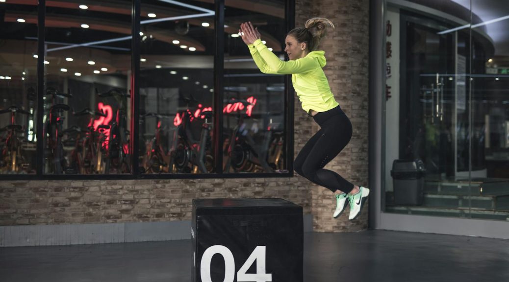 Fit young woman in sports wear executing a box jump indoors, promoting fitness and health in a modern urban gym.